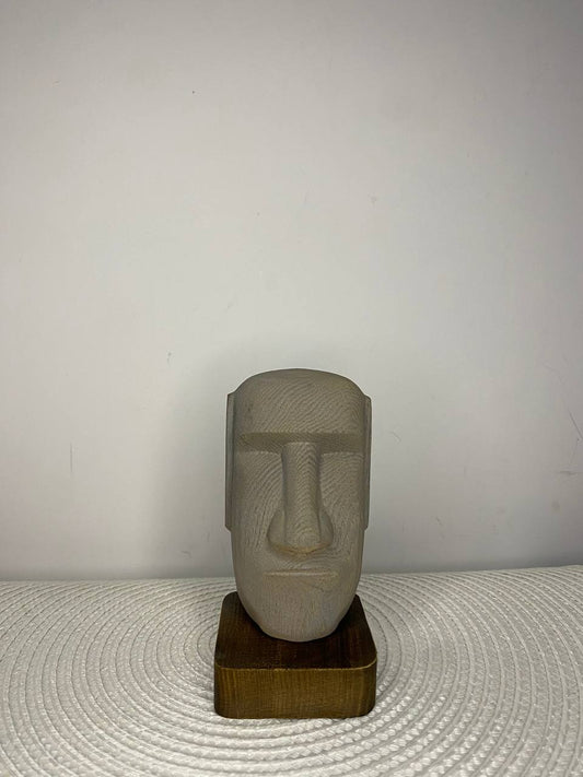 Hand-carved wooden Moai statue inspired by the monumental Easter Island heads, featuring a minimalist elongated face with prominent nose and brow. Crafted from natural ash wood and displayed on a wooden base, the sculpture symbolizes ancestral guardianship, wisdom, resilience, cultural heritage, and enduring strength.