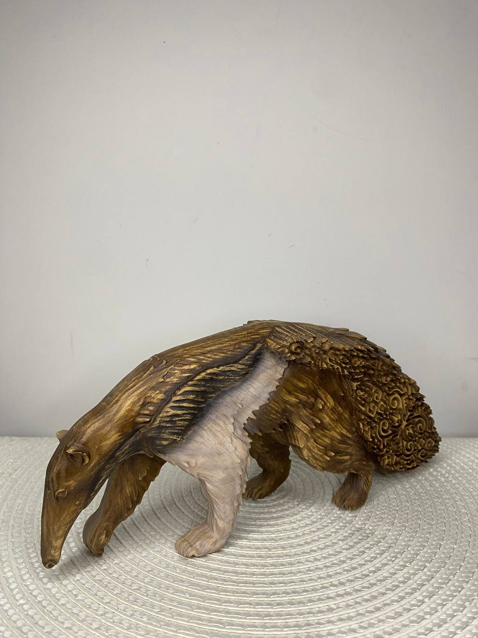 A handcrafted wooden anteater figurine shown in side profile with its head lowered as if searching the ground. The sculpture features a realistic elongated snout, carved fur texture, and a large bushy tail decorated with intricate Celtic knotwork patterns. The body has a warm brown wood finish with lighter natural wood tones on the shoulder and front leg. The figurine stands on a round woven white placemat against a plain light-colored wall.