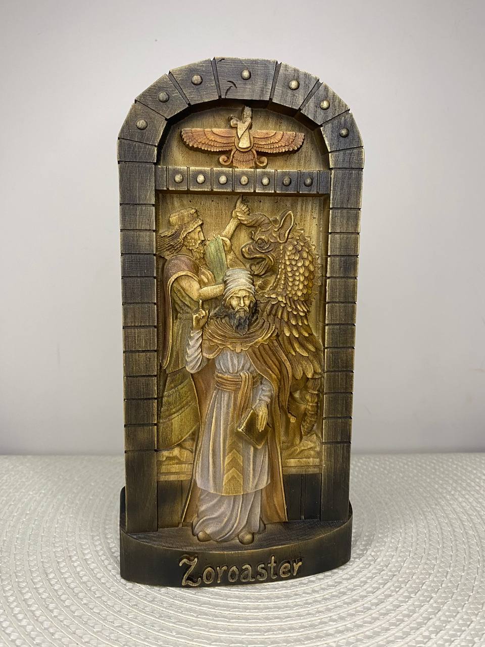 A handcrafted wooden Zoroastrian-inspired relief sculpture displayed within an arched architectural frame finished in dark stained wood with decorative rivet details. At the top of the arch is the Faravahar, the iconic winged symbol of Zoroastrianism. The central scene features a robed priest holding a sacred book, standing before two detailed figures, including a majestic winged creature with finely carved feathers. The artwork is finished in warm brown and golden wood tones with subtle hand-painted highli
