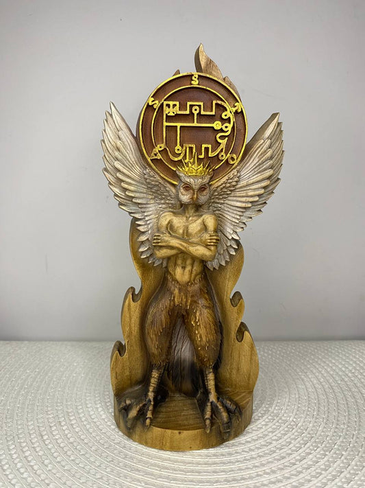 A handcrafted wooden sculpture depicting a winged owl-like humanoid standing with its arms crossed in a commanding pose. The mythical figure features an owl's face with large piercing eyes, feathered wings fully spread, taloned bird feet, and a muscular human-like torso. Above the figure is a circular emblem engraved with an intricate occult sigil, framed by stylized flames. The sculpture is carved from wood with warm golden-brown tones that highlight the detailed feathers, anatomy, and symbo
