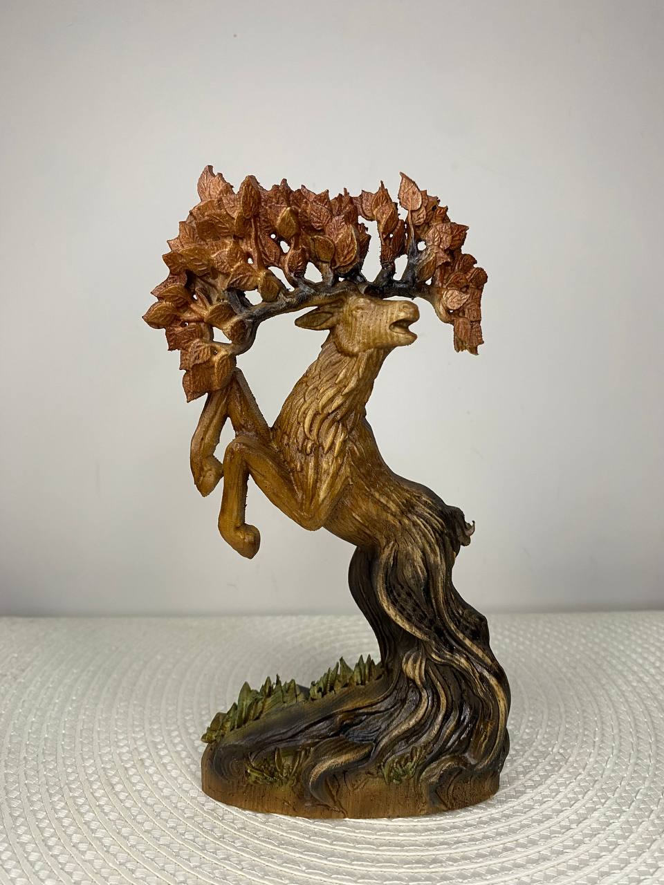 A handcrafted wooden Tree Deer sculpture featuring a rearing deer with a leafy tree canopy growing from its antlers. The detailed carving has flowing wood-grain textures, a twisted tree trunk base, and natural brown and green finishes. Displayed on a white woven placemat against a plain light-colored wall.