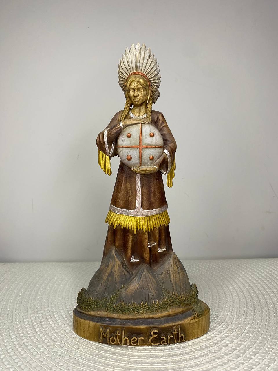 A handcrafted wooden Mother Earth Goddess sculpture depicting a serene woman wearing a feathered headdress and holding a sacred globe. The figure stands on a mountain-inspired base with natural brown, gold, and white finishes, and the base is engraved with the words "Mother Earth." Displayed on a white woven placemat against a plain light-colored wall.