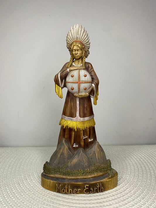A handcrafted wooden Mother Earth Goddess sculpture depicting a serene woman wearing a feathered headdress and holding a sacred globe. The figure stands on a mountain-inspired base with natural brown, gold, and white finishes, and the base is engraved with the words "Mother Earth." Displayed on a white woven placemat against a plain light-colored wall.