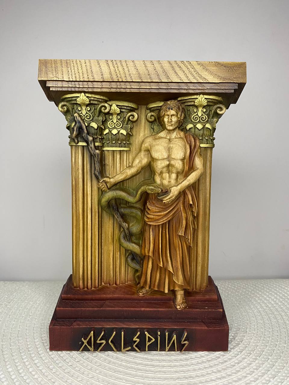 A handcrafted wooden Asclepius sculpture depicting the Greek god of healing standing between classical columns while holding a staff entwined with a serpent. The detailed carving features warm natural wood tones with hand-painted accents, and the base is engraved with the name "Asclepius." Displayed on a white woven placemat against a plain light-colored wall.