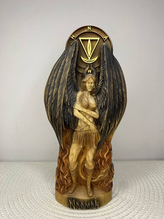 A handcrafted wooden Naamah sculpture depicting a winged female figure standing amid stylized flames with large black feathered wings partially wrapped around her. A symbolic sigil is carved above her, and the base is engraved with the name "Naamah." Finished in warm brown, gold, and black tones, the sculpture is displayed on a white woven placemat against a plain light-colored wall.