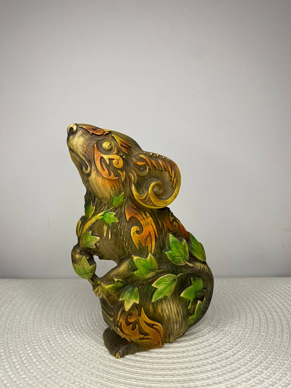 A handcrafted wooden mouse sculpture featuring intricate swirling patterns and vibrant green ivy leaves wrapped around its body. The detailed carving is finished in warm brown, gold, and green tones, with the mouse gazing upward in a lifelike pose. Displayed on a white woven placemat against a plain light-colored wall.