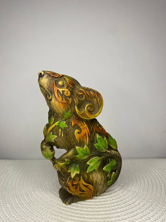 A handcrafted wooden mouse sculpture featuring intricate swirling patterns and vibrant green ivy leaves wrapped around its body. The detailed carving is finished in warm brown, gold, and green tones, with the mouse gazing upward in a lifelike pose. Displayed on a white woven placemat against a plain light-colored wall.
