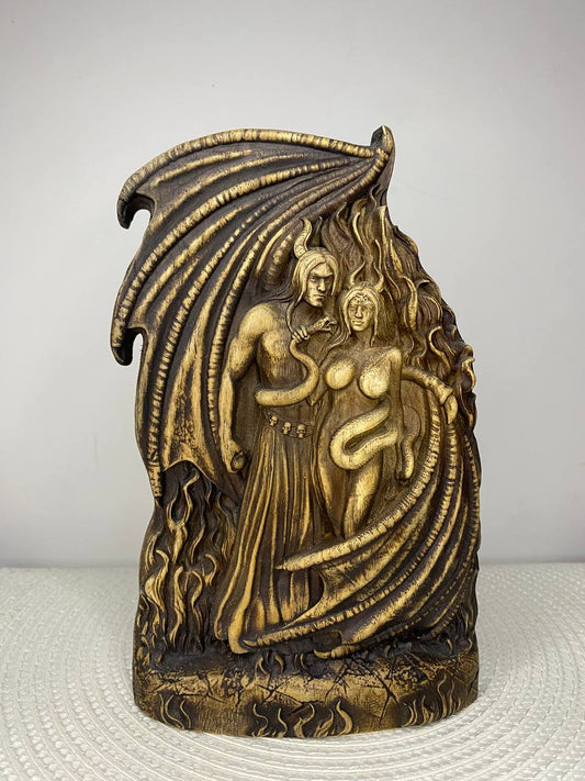 A handcrafted wooden sculpture depicting Lucifer and Lilith standing together beneath a large bat-like wing. A serpent coils around Lilith while flames surround the figures, creating a dramatic gothic design. The detailed carving is finished in warm brown and black tones and is displayed on a white woven placemat against a plain light-colored wall.