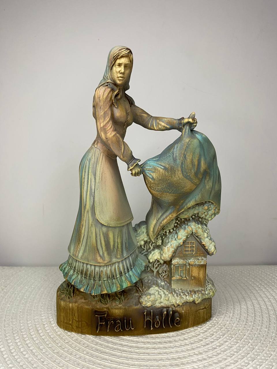 A handcrafted wooden Frau Holle sculpture depicting the legendary German fairy tale figure shaking out a large blanket above a snow-covered cottage. The detailed carving features soft blue, white, and natural wood tones, with the base engraved "Frau Holle." Displayed on a white woven placemat against a plain light-colored wall.