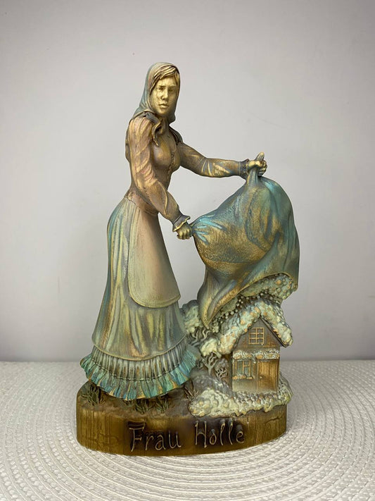 A handcrafted wooden Frau Holle sculpture depicting the legendary German fairy tale figure shaking out a large blanket above a snow-covered cottage. The detailed carving features soft blue, white, and natural wood tones, with the base engraved "Frau Holle." Displayed on a white woven placemat against a plain light-colored wall.