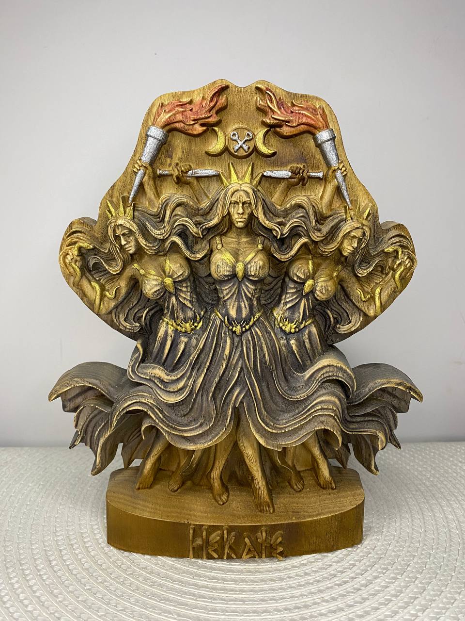 A handcrafted wooden Hecate sculpture depicting the triple goddess with three female figures standing back-to-back beneath crossed torches, twin crescent moons, and crossed daggers. The detailed carving is finished in warm gold and dark brown tones, with the base engraved "Hecate." Displayed on a white woven placemat against a plain light-colored wall.