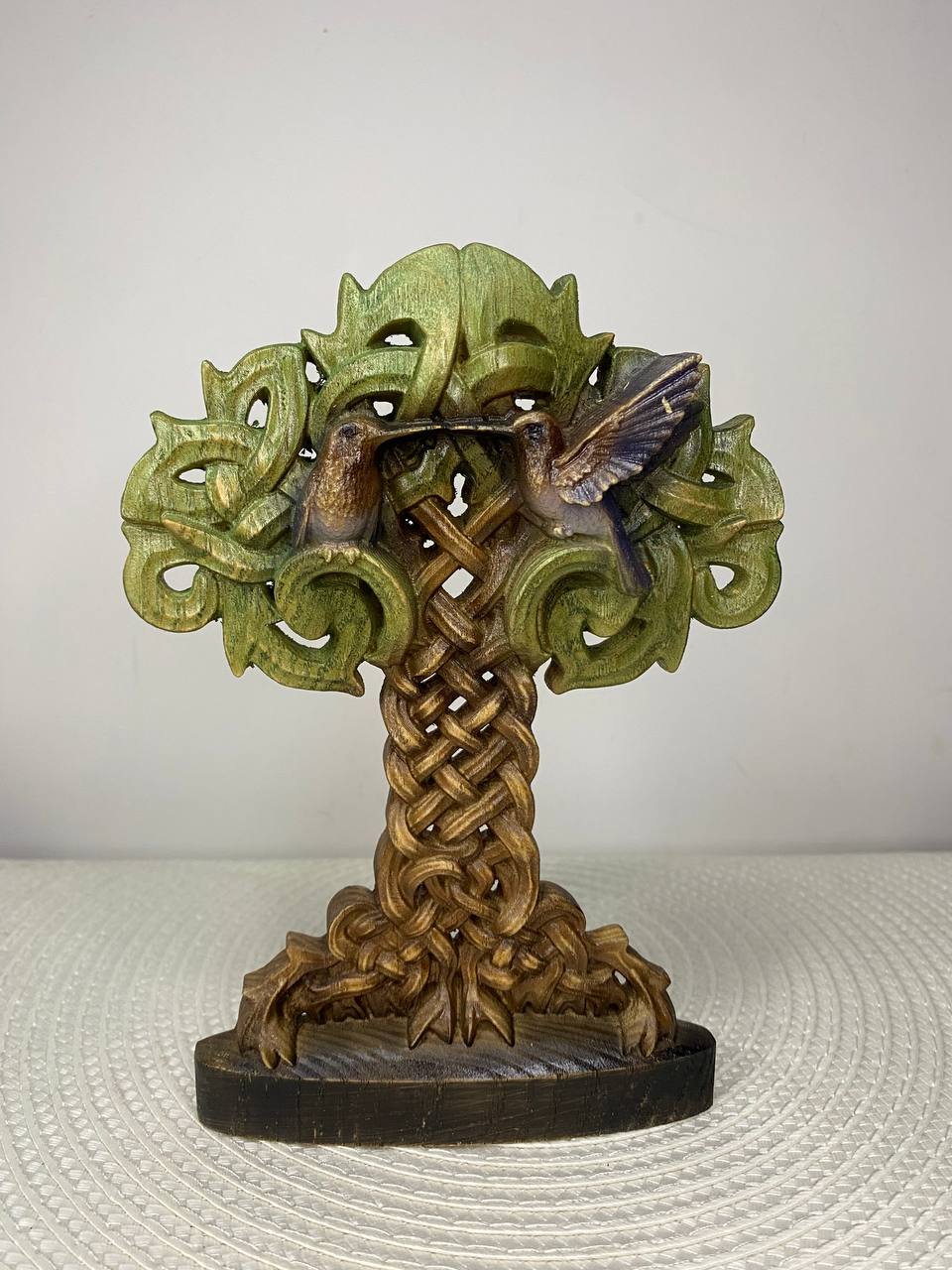 A decorative carved tree sculpture with two hummingbirds in flight, featuring a green leafy canopy, a braided brown trunk, and a dark oval base, photographed against a plain light background.