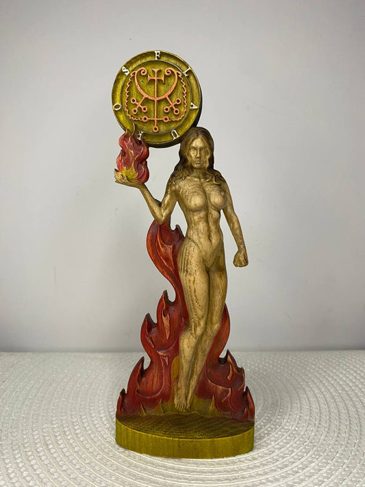 A handcrafted wooden Flauros sculpture depicting a female figure emerging from stylized flames while holding a blazing fire in one hand. An ornate occult sigil is carved above her, and the sculpture is finished in warm natural wood tones with striking red and gold accents. Displayed on a white woven placemat against a plain light-colored wall.