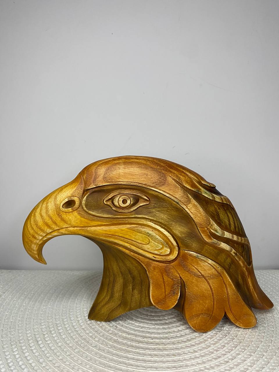 A handcrafted wooden eagle head sculpture featuring a bold side profile with a sharply curved beak and detailed carved eye. The minimalist design highlights the natural wood grain in warm golden-brown tones with subtle darker accents. Displayed on a white woven placemat against a plain light-colored wall.