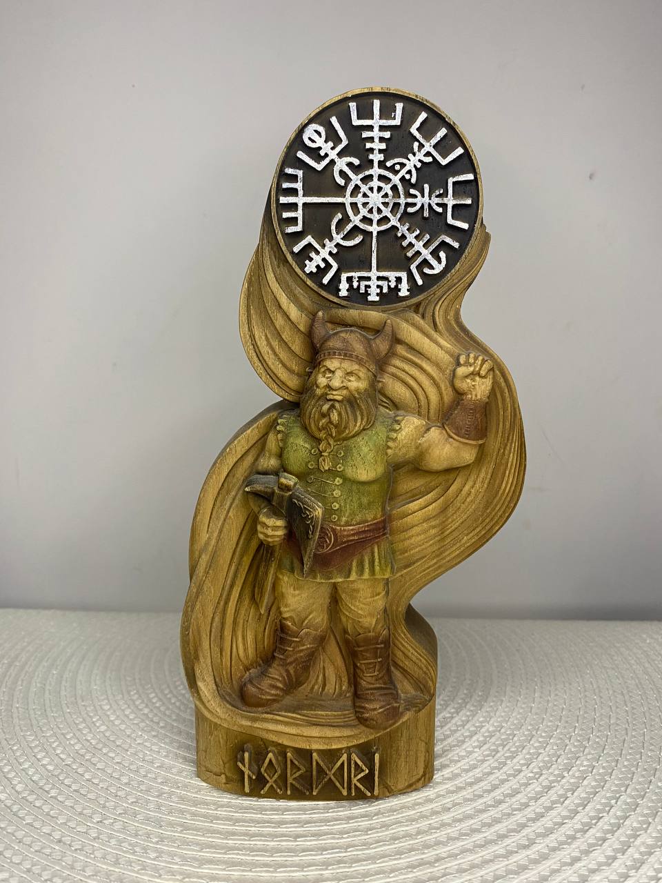 A carved wooden Viking warrior with a long beard, helmet, axe, and raised hand, standing beneath a circular Norse symbol. The figure is mounted on a decorated wooden base with runic characters and displayed against a plain light-colored background.