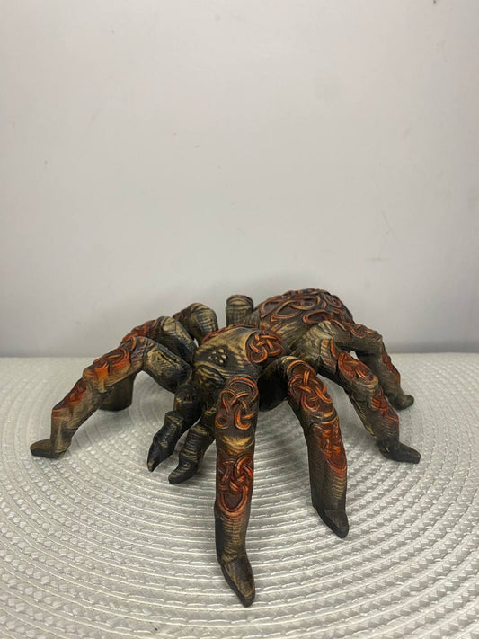 Alt text: Hand-carved wooden spider figurine with a dark, rustic finish and eight long, curved legs. Intricate Celtic knotwork and swirling decorative patterns cover the spider’s body and legs, creating a detailed Nordic-inspired design. The sculpture is displayed on a textured white surface against a plain light-colored background.