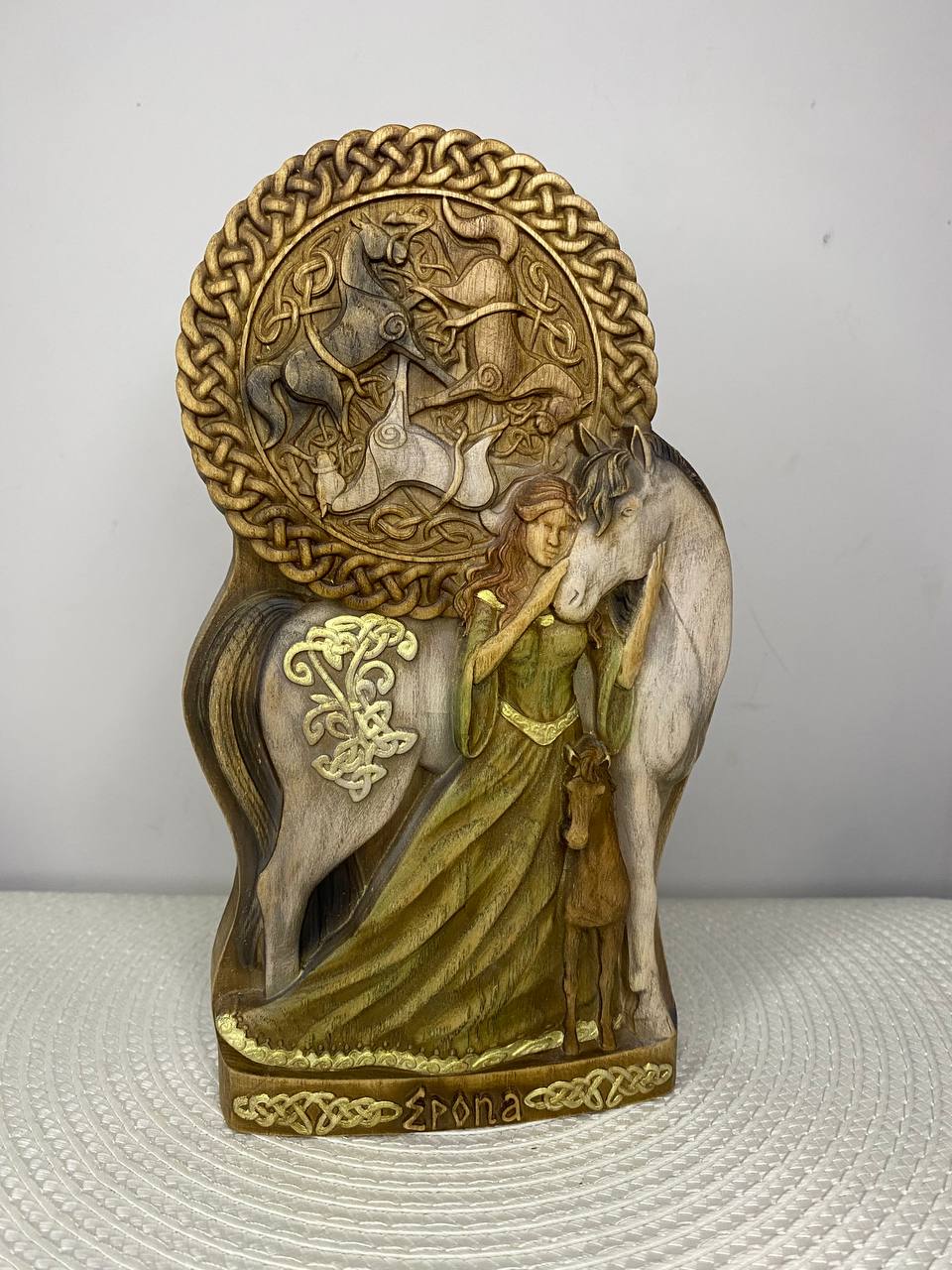 Hand-carved wooden statue of Epona, the Celtic horse goddess, standing between two horses with a flowing green robe. A large circular Celtic knotwork panel featuring horses rises behind her, with intricate carvings and decorative knotwork throughout the sculpture.