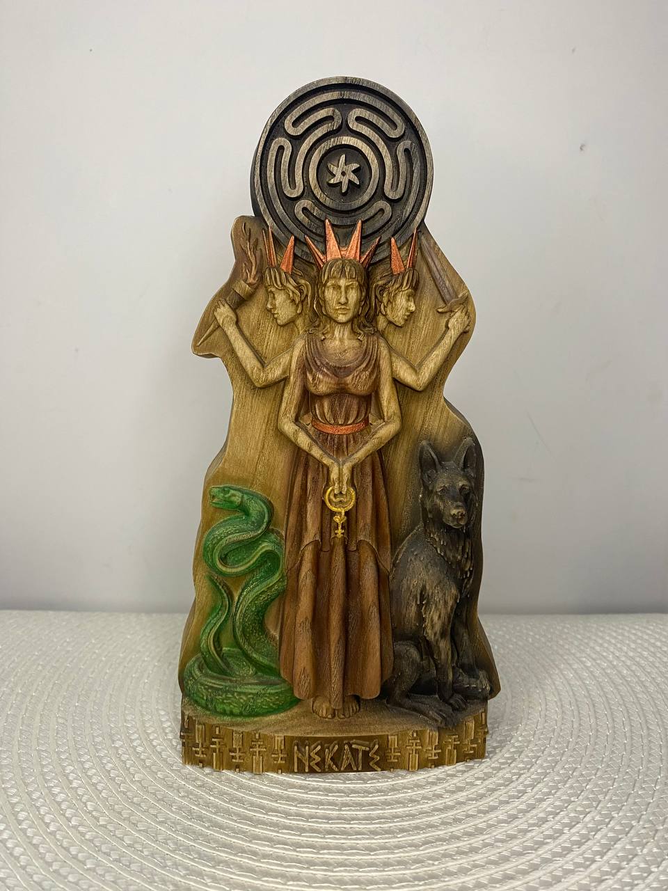 Hand-carved wooden statue of Hecate, the Greek goddess of witchcraft and crossroads, depicted as a three-faced figure in a long robe. She stands beside a green serpent and a black dog, with torches behind her and a circular symbol above. The base is decorated with carved Greek-inspired symbols.