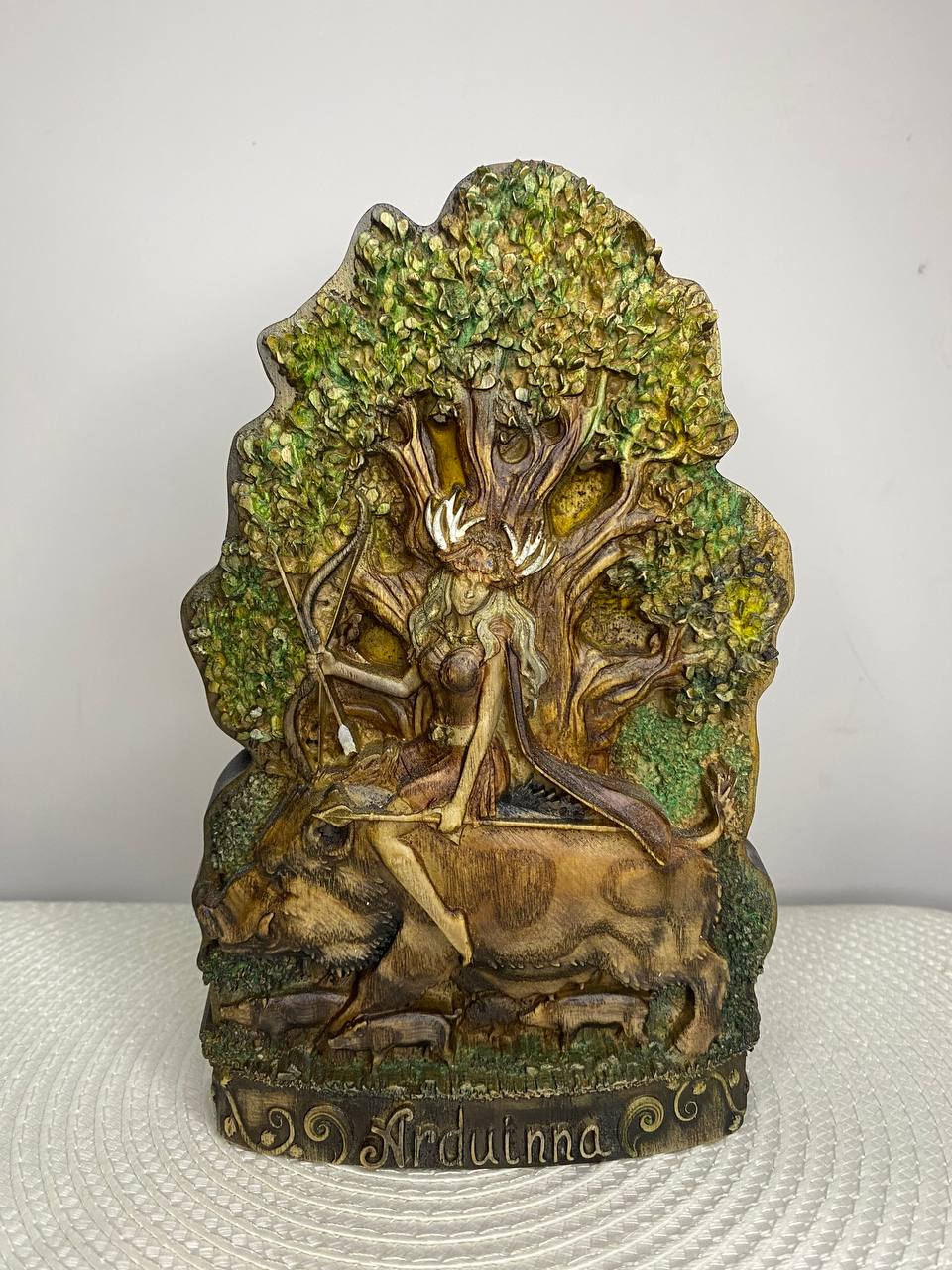 Hand-carved wooden statue of Arduinna, the Celtic goddess of the forest and hunting, depicted as a bearded figure with antlers riding a wild boar beneath a large leafy tree. The detailed sculpture features natural woodland elements, green foliage, and a decorative base engraved with “Arduinna.”