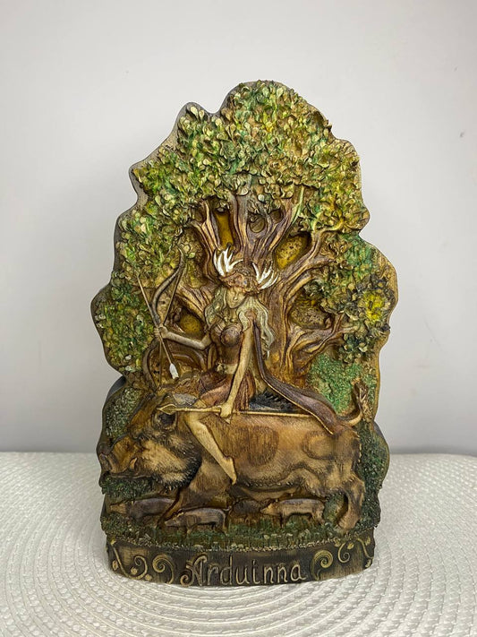 Hand-carved wooden statue of Arduinna, the Celtic goddess of the forest and hunting, depicted as a bearded figure with antlers riding a wild boar beneath a large leafy tree. The detailed sculpture features natural woodland elements, green foliage, and a decorative base engraved with “Arduinna.”