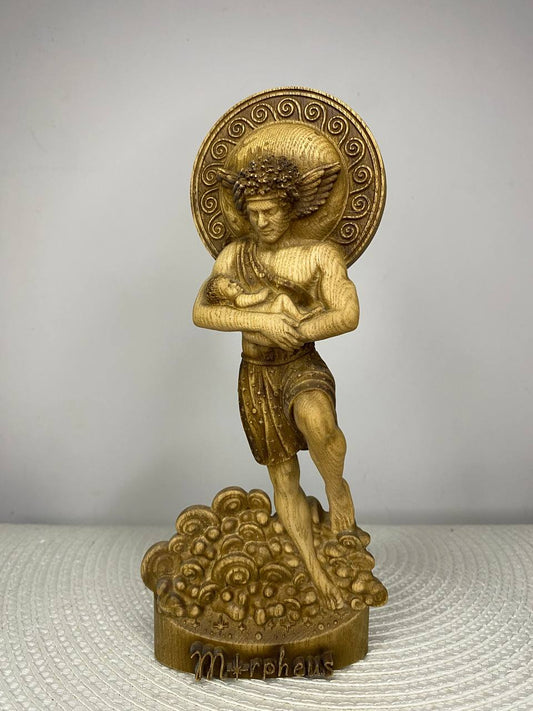 Hand-carved wooden statue of Morpheus, Greek god of dreams, holding a sleeping infant in his arms while standing upon stylized clouds. The figure features delicate wings and a circular ornamental halo, symbolizing peaceful sleep, protection, innocence, divine guidance, and dream-inspired mythology decor.
