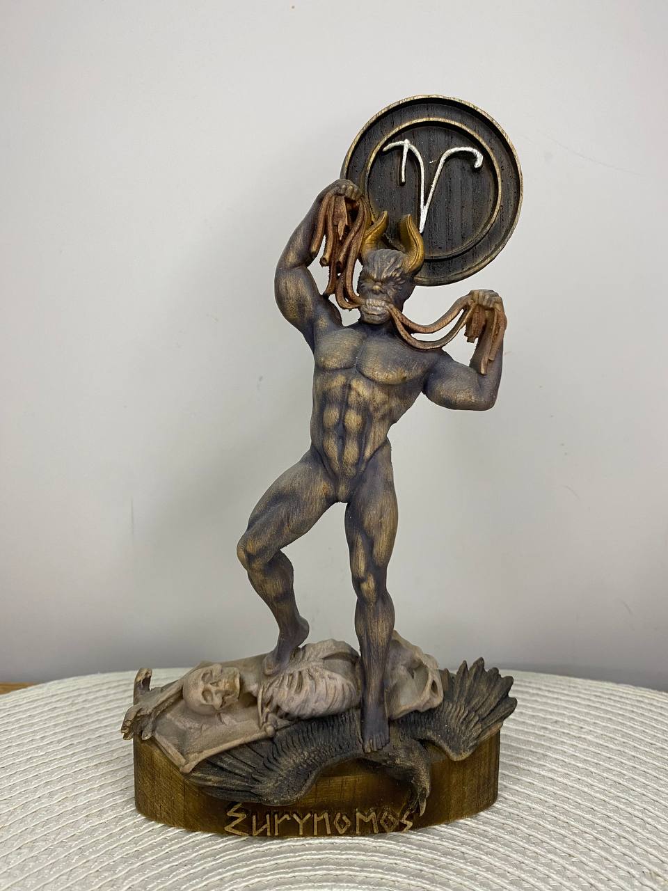 Handcrafted wooden Eurynomous statue inspired by Greek underworld mythology, featuring the fearsome demon standing over a skeleton with ram horns, an Aries zodiac symbol, and hand-painted natural wood, bronze, and gray tones. Mythology altar decor and collectible sculpture.