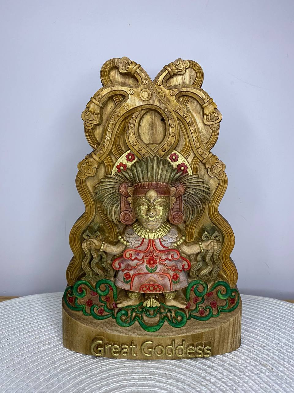 Handcrafted wooden Great Goddess statue inspired by ancient Mesoamerican mythology, featuring an ornate headdress, ceremonial attire, sacred serpent motifs, and an elaborate temple-style backdrop, hand-painted in natural wood, gold, green, and red tones. Mythology altar decor and collectible sculpture.