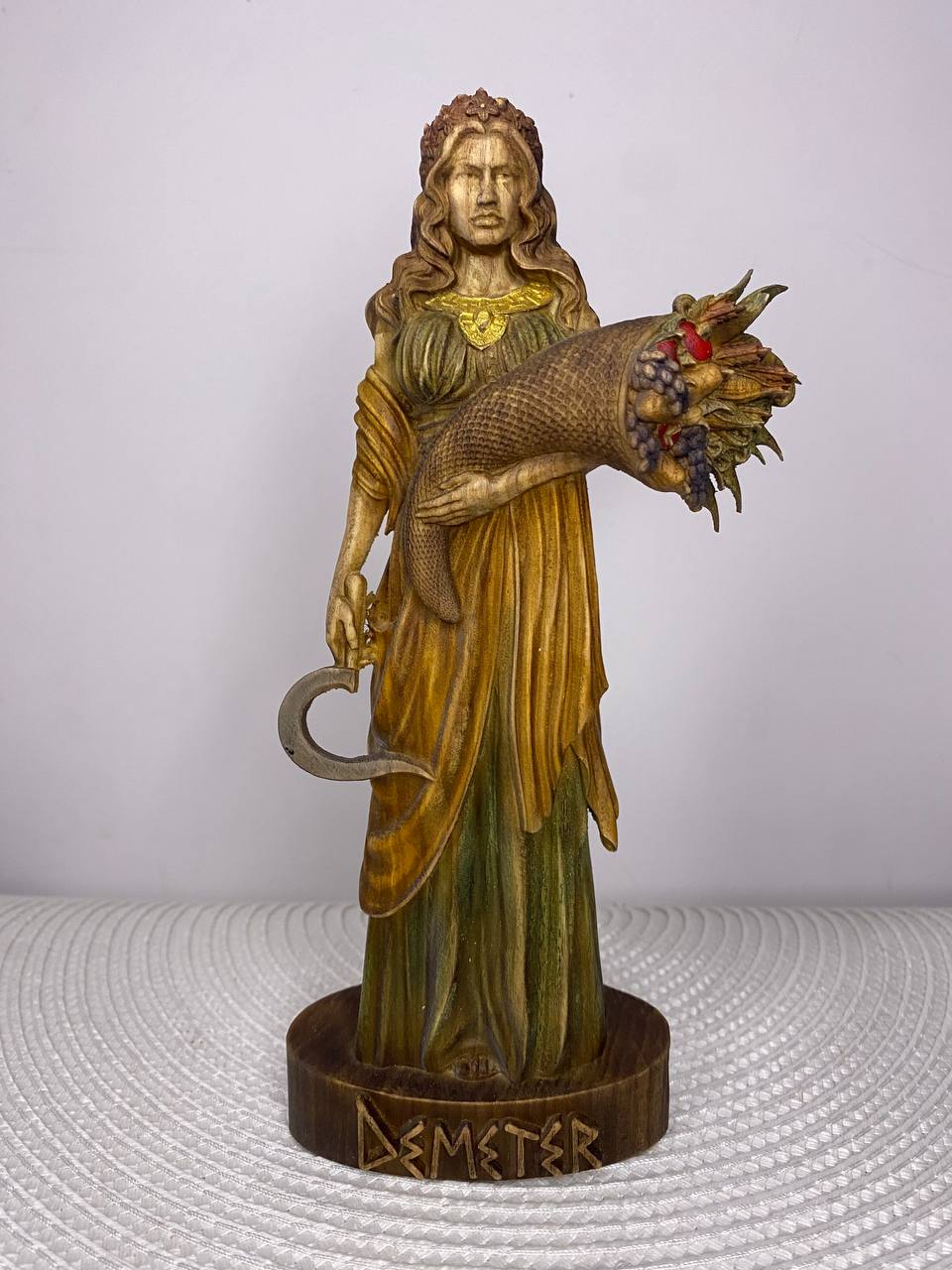 Handcrafted wooden Demeter statue inspired by the Greek goddess of agriculture and harvest, holding a cornucopia overflowing with fruits and grain and a traditional sickle, hand-painted in natural wood, green, gold, and earthy tones. Greek mythology altar decor and collectible sculpture.